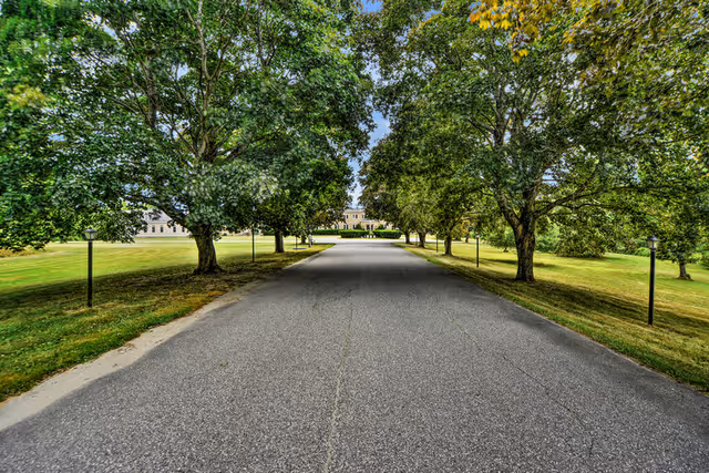 A paved driveway lined with green trees on both sides leading to a large building in the distance under a partly cloudy sky.