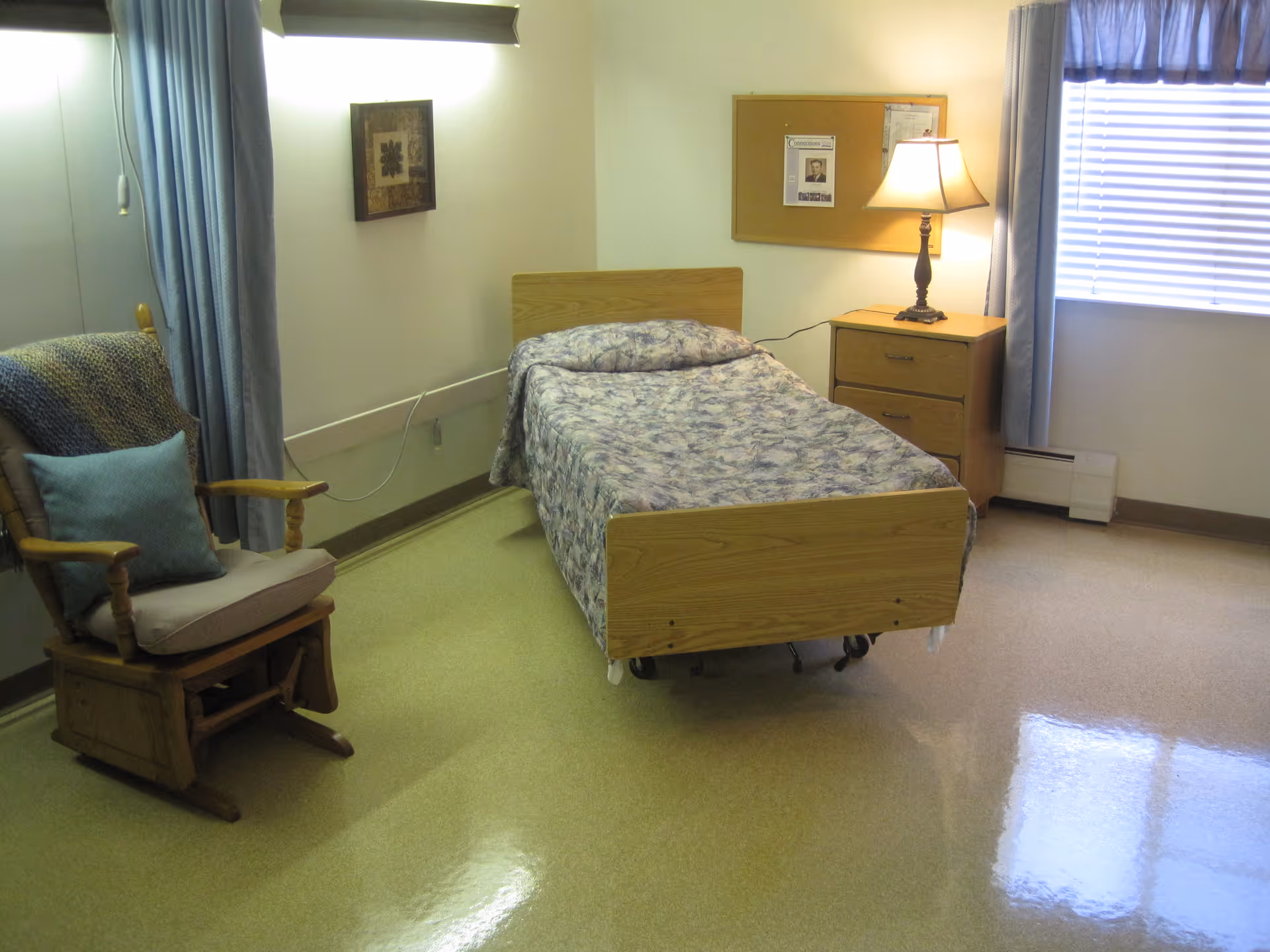 A simple bedroom with a single bed covered in a floral bedspread, a wooden rocking chair with cushions, a wooden nightstand with a lamp, a bulletin board on the wall, and a window with blue curtains and blinds.