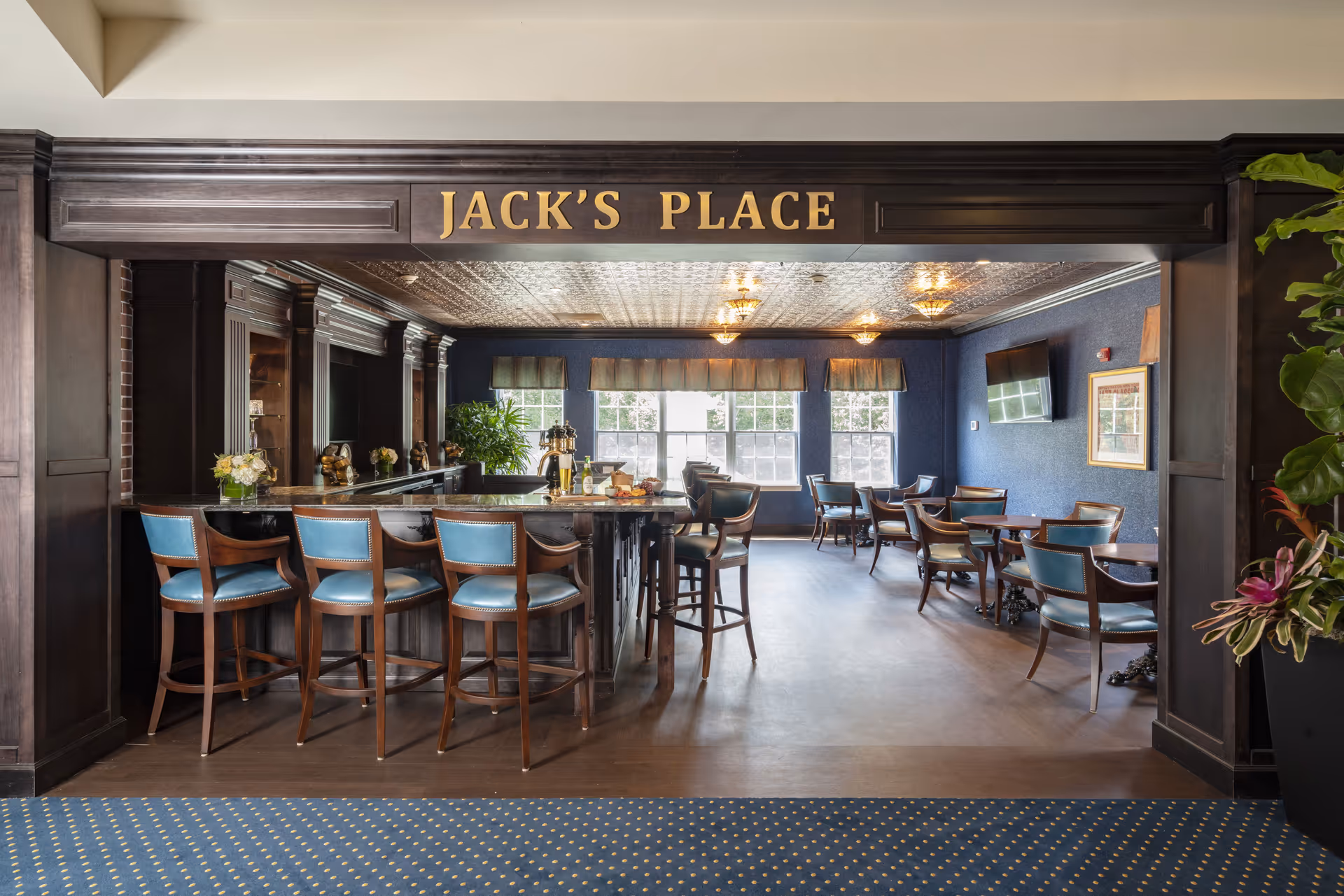 Interior view of a lounge area named 'Jack's Place' featuring a bar with high wooden chairs upholstered in blue, several small tables with matching chairs, large windows with valances, dark wood paneling, and decorative ceiling lights.