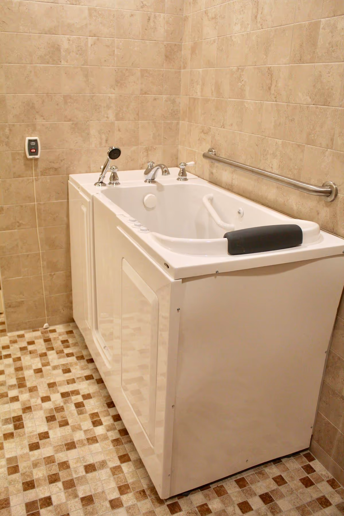 A walk-in bathtub with a built-in seat and handrails, surrounded by beige tiled walls and a checkered tile floor. The bathtub has a handheld showerhead, faucet controls, and a black headrest.