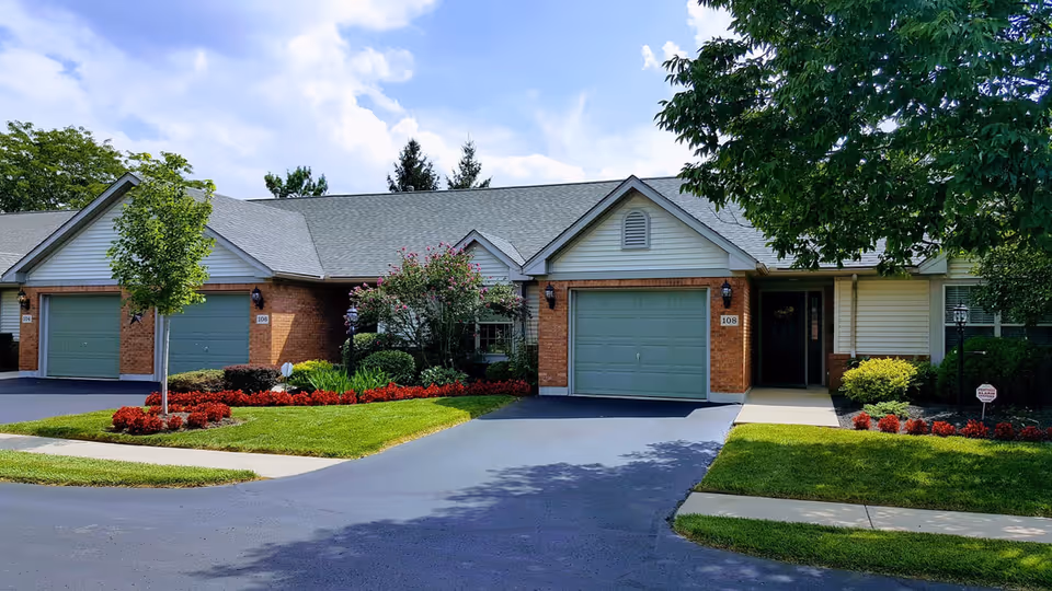 Exterior view of a single-story senior living facility building with attached garages, well-maintained landscaping including green lawns, red flowers, and trees under a partly cloudy sky.