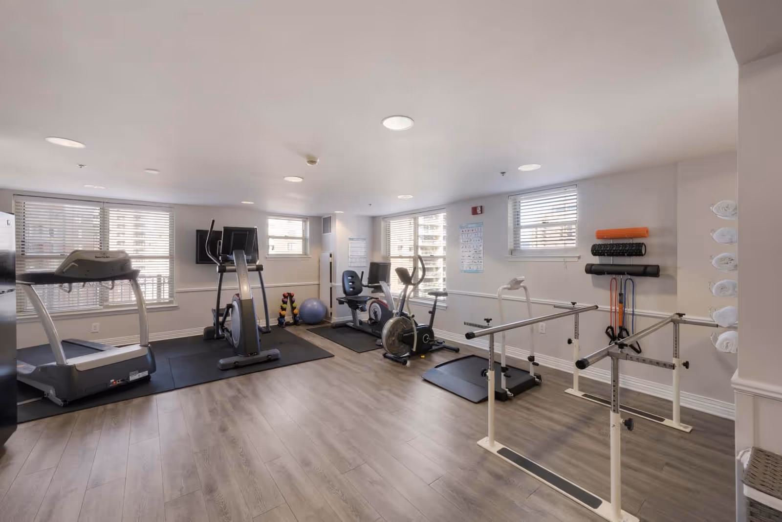 A bright exercise room with large windows and light wood flooring. The room contains various fitness equipment including a treadmill, elliptical machine, stationary bike, hand weights, exercise balls, resistance bands, foam rollers, and parallel bars for balance and rehabilitation exercises. White towels are neatly rolled and stored on a rack on the right side.