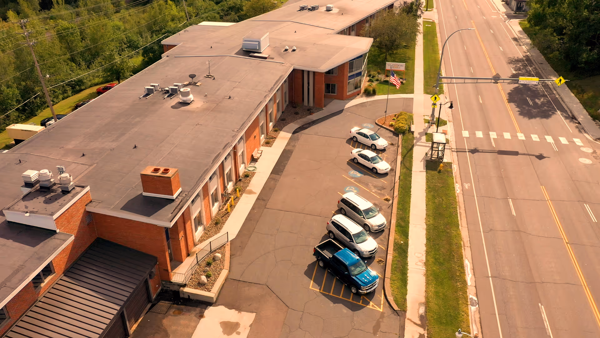 Aerial view of a brick senior living building with a small parking lot and cars next to a main road.