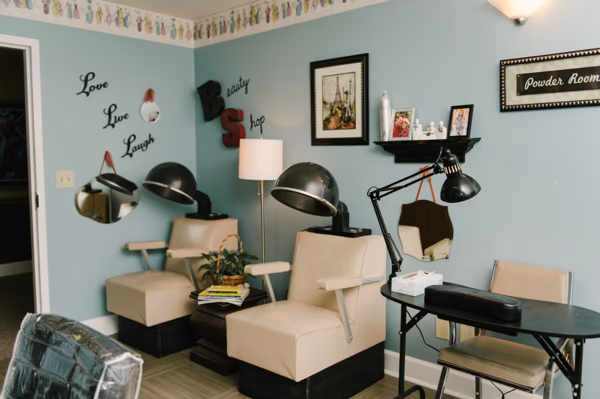 Interior of a beauty shop area with two beige salon chairs under vintage hair dryers, a small table with magazines and a plant, a black manicure table with a lamp and chair, light blue walls decorated with framed pictures, mirrors, and wall art including the words 'Love Live Laugh' and 'Powder Room'.