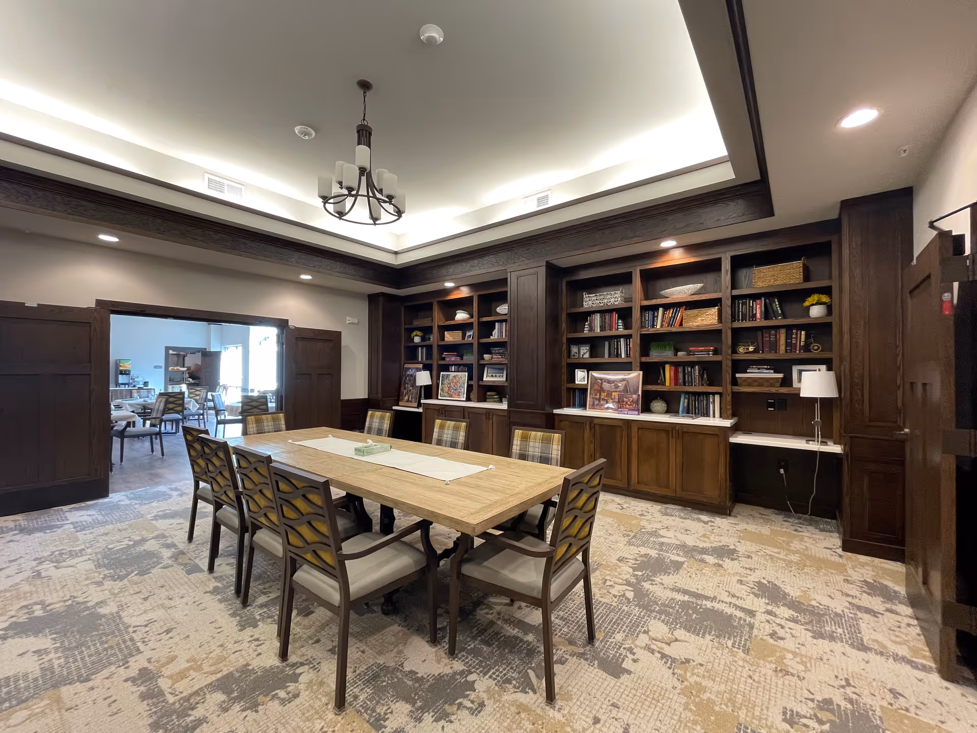 A well-lit interior room with a long wooden table surrounded by eight chairs with patterned cushions. The room features built-in dark wood bookshelves filled with books, baskets, and decorative items. A chandelier hangs from the ceiling, and there is a doorway leading to another room with additional seating and tables.
