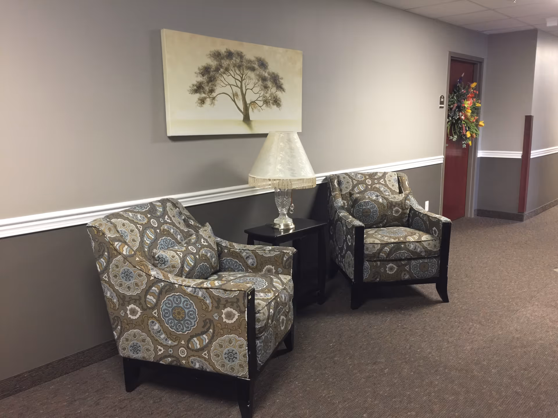 A hallway seating area in a retirement community featuring two patterned armchairs with matching cushions, a small black side table with a decorative lamp, and a wall painting of a tree. The hallway has beige and brown walls with white trim and a door decorated with a colorful floral wreath.