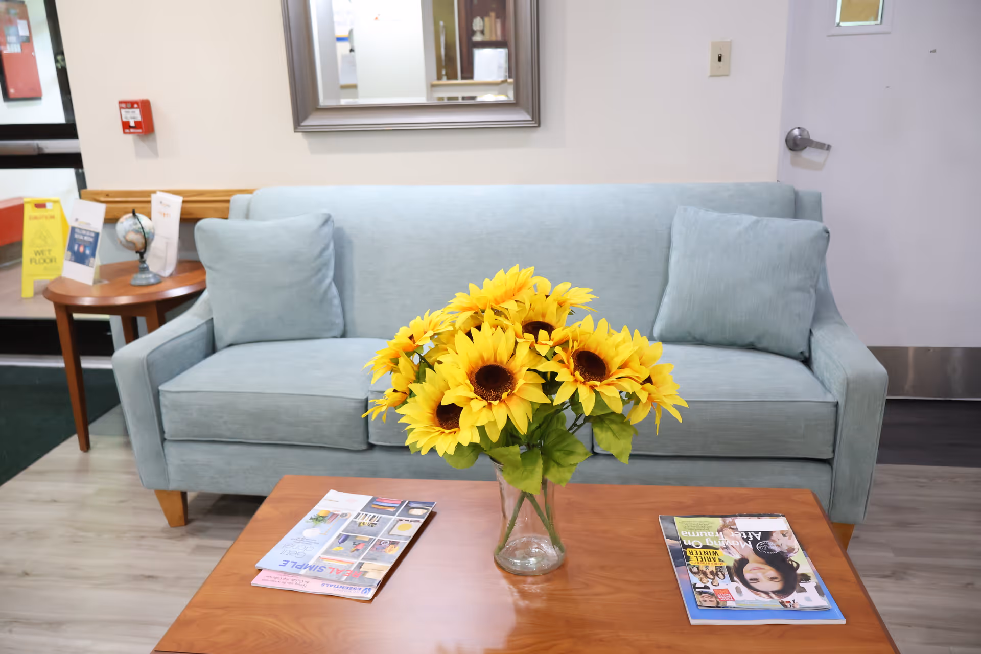 A light blue sofa with two matching cushions in a waiting area. In front of the sofa is a wooden coffee table with a vase of bright yellow sunflowers and two magazines. To the left, there is a small round wooden side table with a small globe and some papers. A mirror hangs on the wall above the sofa, and a door is visible on the right side of the image. A yellow caution wet floor sign is seen near the entrance in the background.