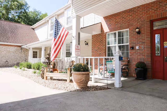 Exterior view of a senior living facility entrance with a white porch railing, an American flag, potted plants, a bench, and a red door. The building has brick and white siding walls, and there is a 'No Parking Loading Zone' sign near the porch.