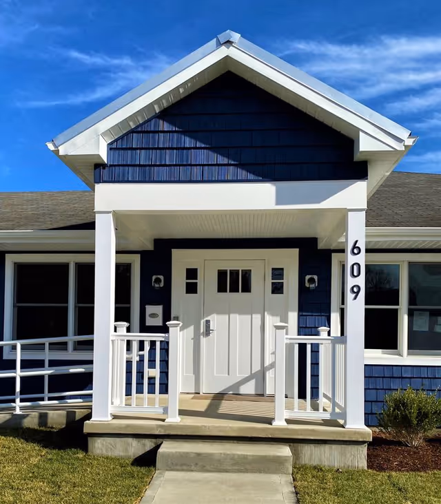 Front entrance of a blue and white building with a small porch, white double doors, and the number 609 displayed vertically on the right pillar. There is a ramp on the left side and a small bush on the right side under a clear blue sky.