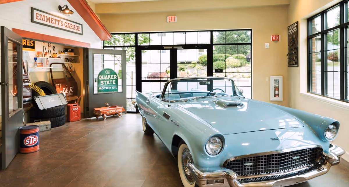 Interior view of a vintage-themed garage area inside a facility, featuring a light blue classic convertible car parked on a tiled floor. To the left, there is a small garage setup labeled 'Emmett's Garage' with various automotive items like tires, oil cans, and a red toy wagon. Large windows and glass doors provide natural light and a view of greenery outside.
