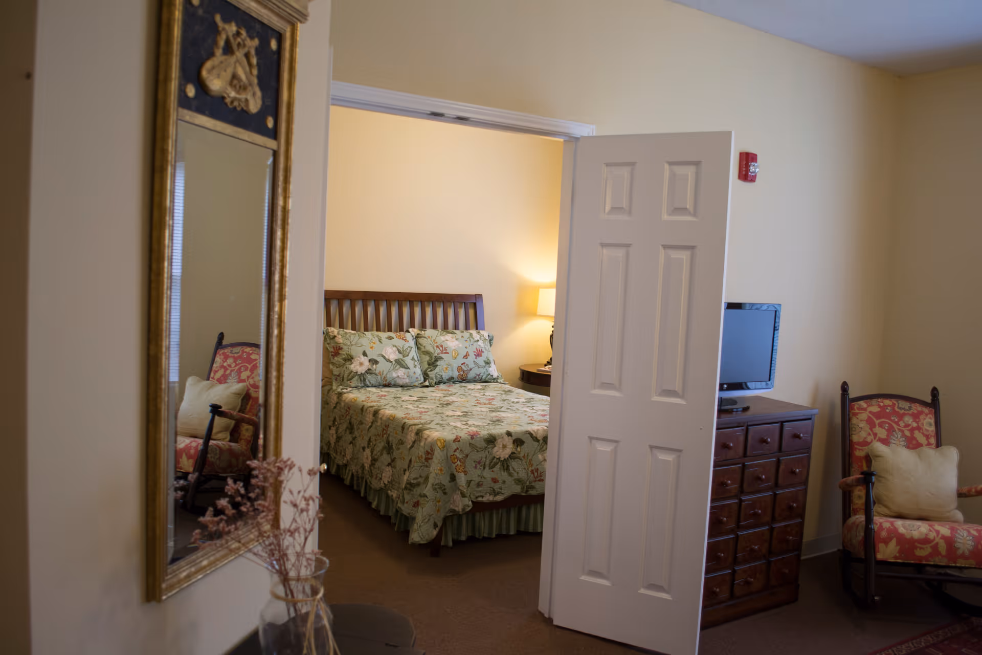 View into a bedroom with a bed covered in a floral bedspread, a nightstand with a lamp, a wooden dresser with a small TV on top, a rocking chair with a cushion, and a decorative mirror on the wall in the foreground.
