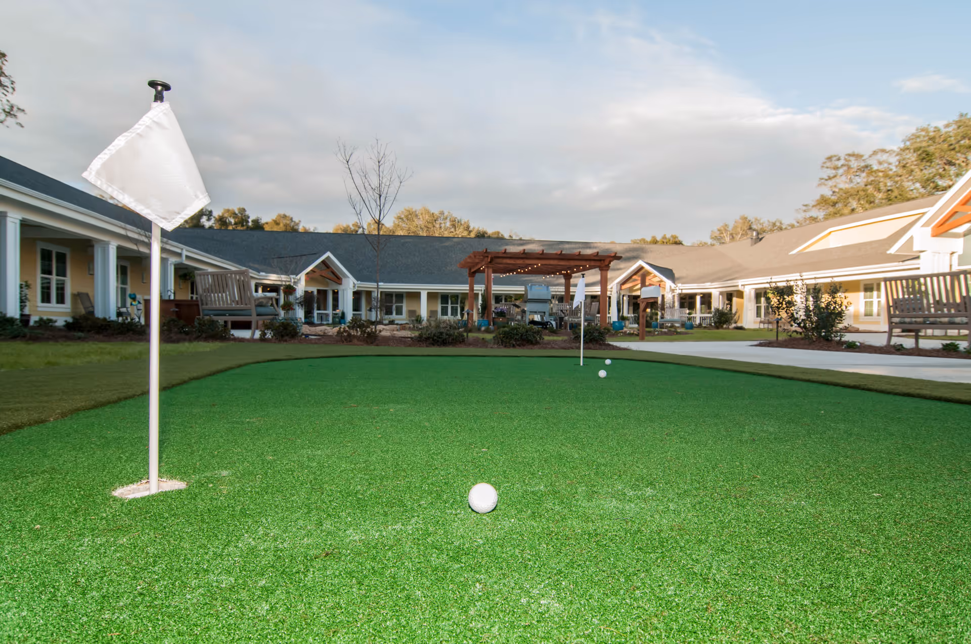 Outdoor putting green with three golf balls and two white flags in front of a senior living facility building with benches, a pergola, and landscaped grounds under a cloudy sky.