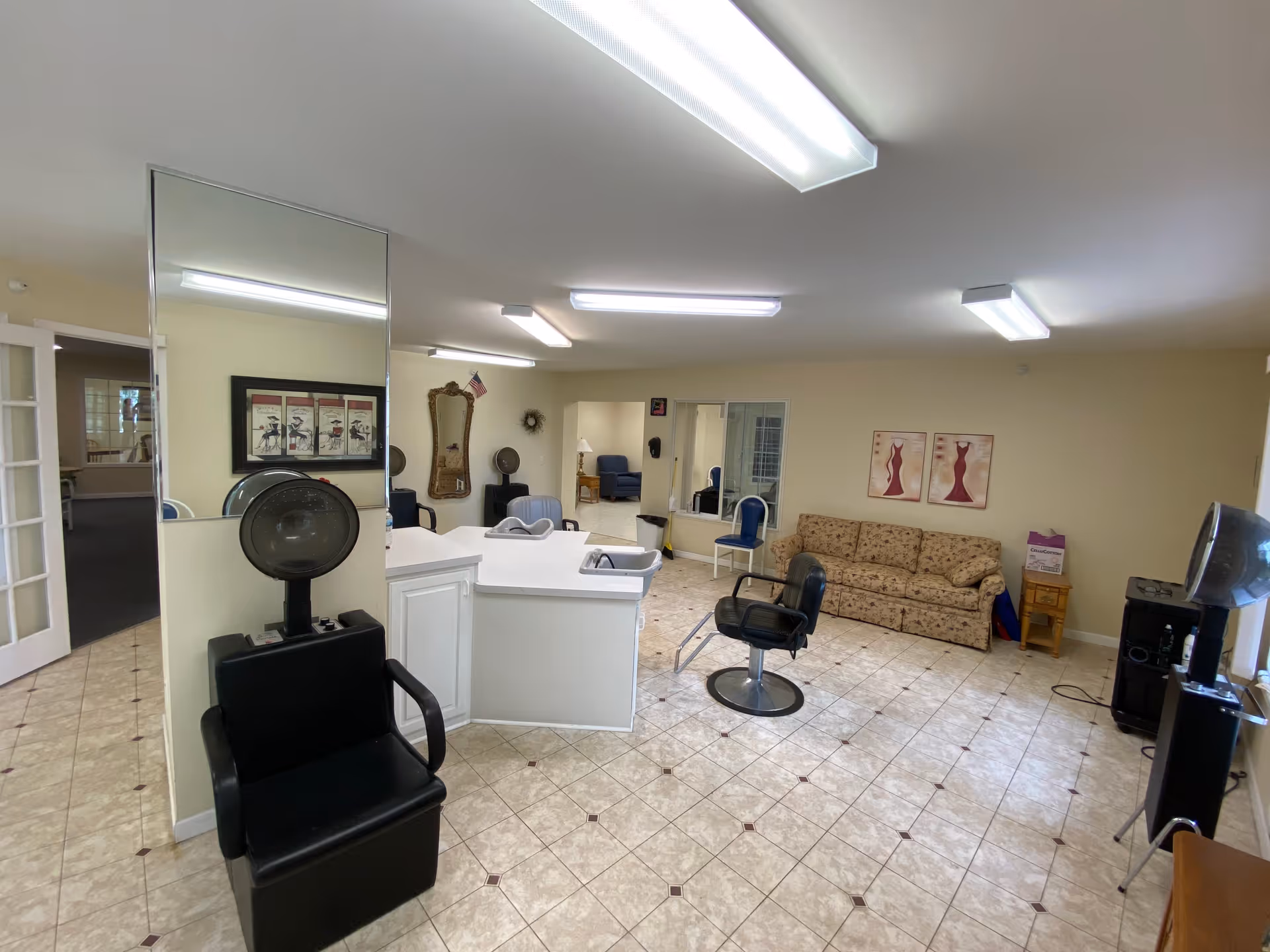 Interior view of a salon area in an assisted living facility with two black salon chairs and hair dryers, a white reception desk, a floral patterned couch, blue chairs, wall decorations including framed pictures and a mirror, and tiled flooring under bright fluorescent lighting.