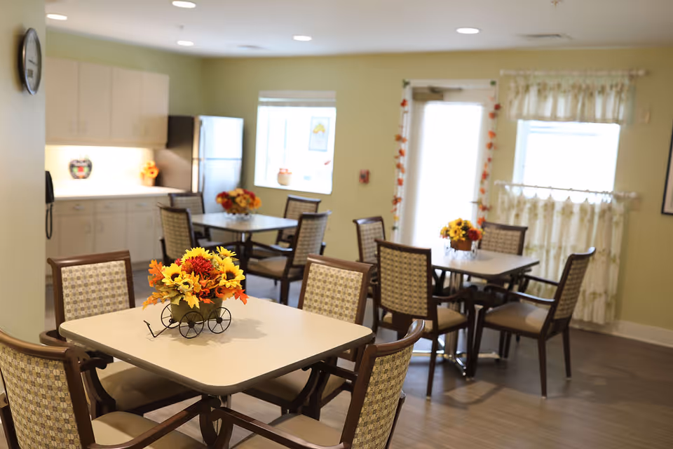 A bright dining room in a senior living facility with several square tables, each surrounded by four cushioned chairs. Each table has a centerpiece of colorful artificial flowers. The room has light green walls, a clock on one wall, a refrigerator, cabinets, and windows with floral curtains letting in natural light.