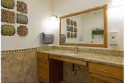 A bathroom vanity area with a large mirror above a granite countertop. The countertop has a built-in sink with a faucet. Below the countertop are wooden cabinets and drawers. On the wall to the left of the mirror, there is a decorative arrangement of square tiles with floral patterns. A paper towel dispenser and a soap dispenser are mounted on the walls near the sink. The walls are light-colored with a tiled backsplash.