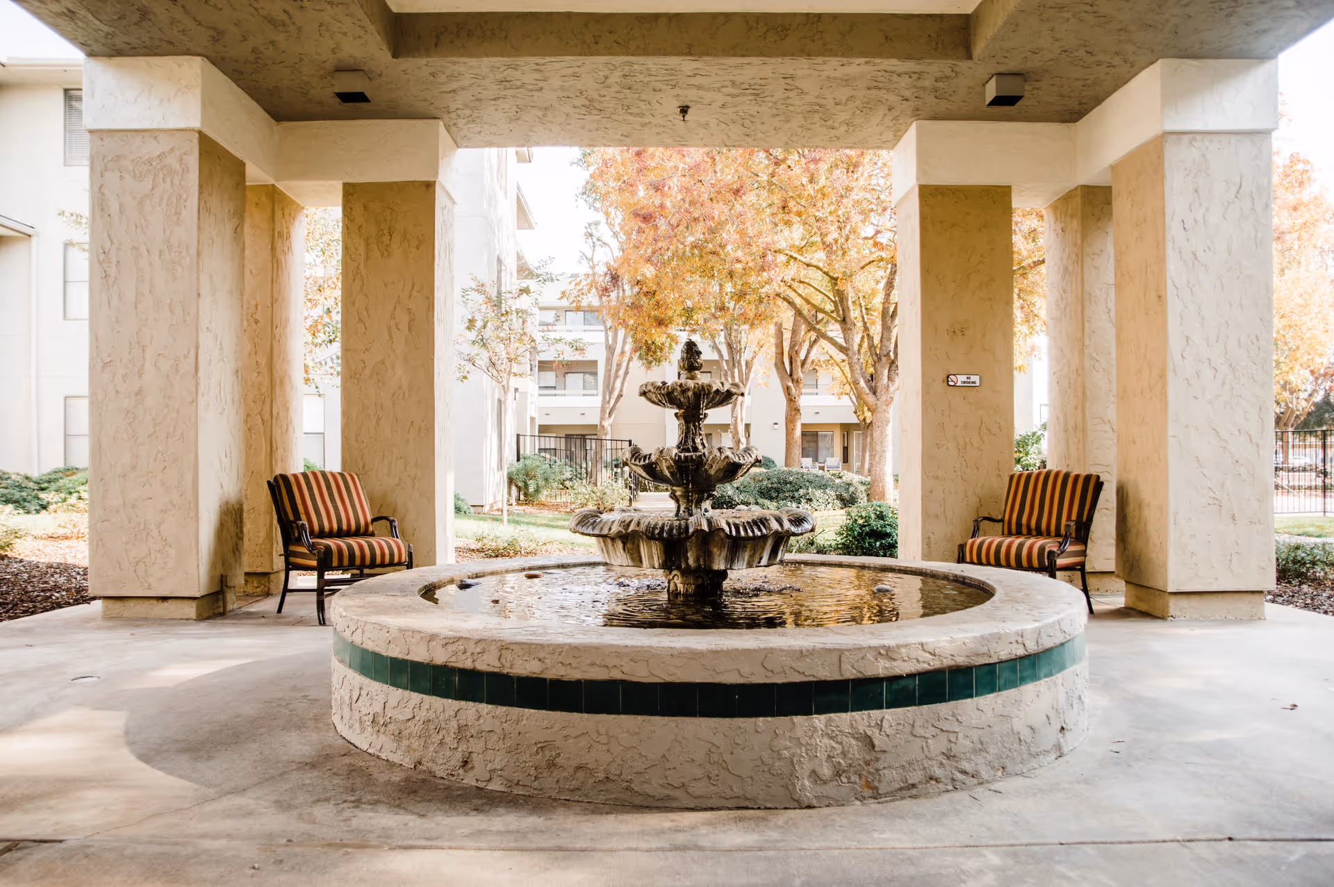 Covered outdoor seating area with a round stone water fountain in the center, flanked by two striped cushioned chairs. The background shows trees with autumn foliage and parts of the building exterior.