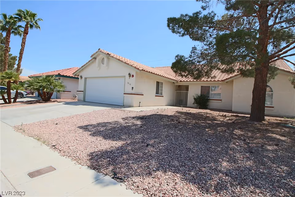 Exterior view of a single-story residential home with a tiled roof, a two-car garage, and a front yard covered with gravel and a large tree casting a shadow. There are palm trees and other greenery visible in the background under a clear blue sky.
