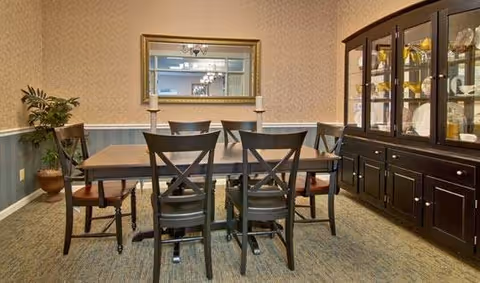 A dining room with a rectangular wooden table surrounded by six dark wooden chairs. On the table are two white candles in holders. Behind the table is a large mirror on the wall reflecting a chandelier. To the right is a dark wooden china cabinet displaying various dishes and glassware. A potted plant is placed in the corner to the left. The walls have a two-tone design with wallpaper on the upper half and blue paneling on the lower half.