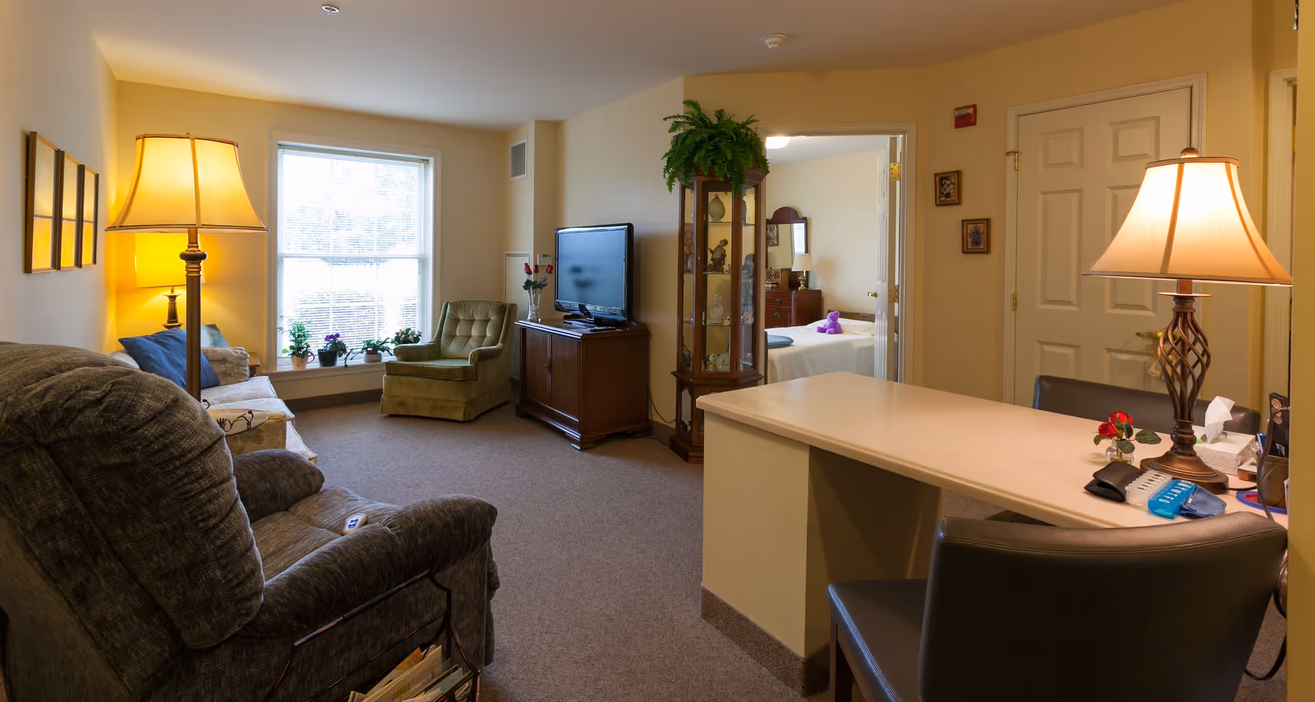 Well-lit living room in a senior facility with armchairs and a sofa, a TV on a cabinet, lamps, a desk area, and a doorway leading to a bedroom.