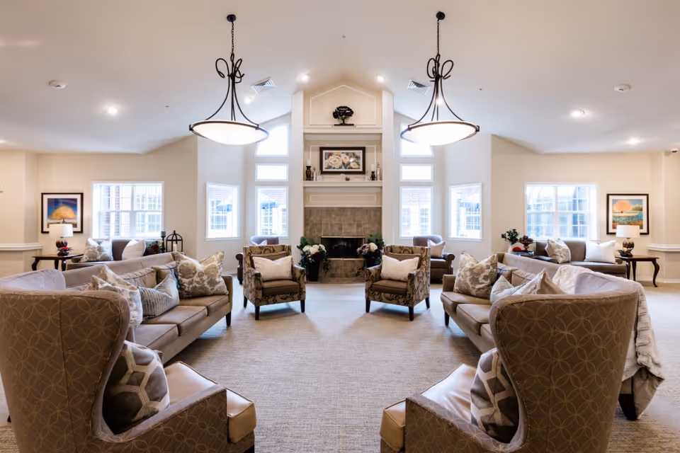 Spacious, well-lit communal living room with sofas, armchairs, chandeliers, and a central fireplace.