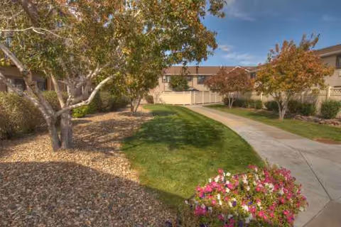 Curved outdoor walkway leads past trees and flowers to a beige senior facility.