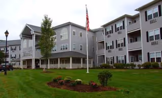 Exterior view of a gray senior living building with a manicured lawn and flag.
