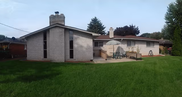 Exterior view of a single-story brick building with a chimney, a small patio area with outdoor chairs and an umbrella, surrounded by a green lawn and trees in the background under a partly cloudy sky.