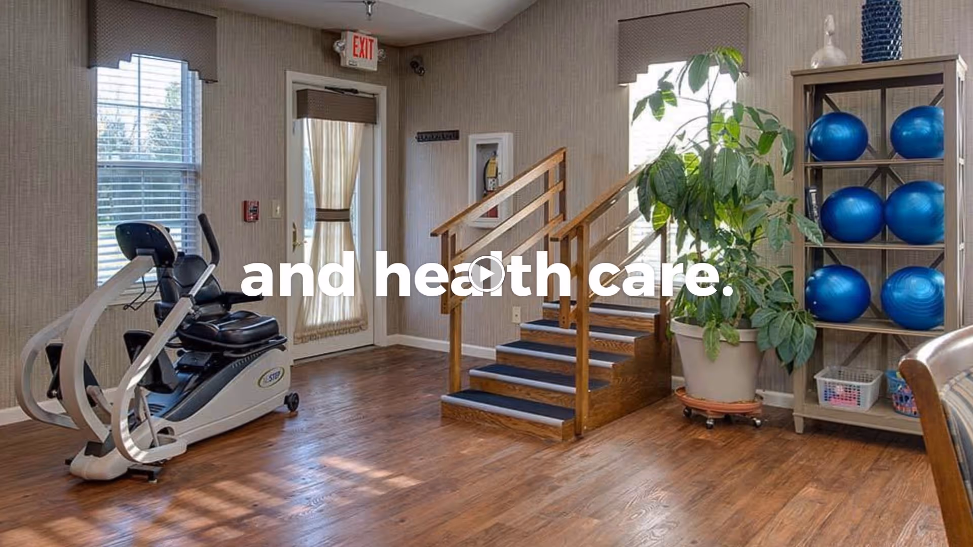A fitness room with wooden flooring featuring an exercise bike, a wooden staircase with handrails, a large potted plant, and a shelving unit holding several blue exercise balls. There are windows with blinds and a door with a curtain, allowing natural light into the room.