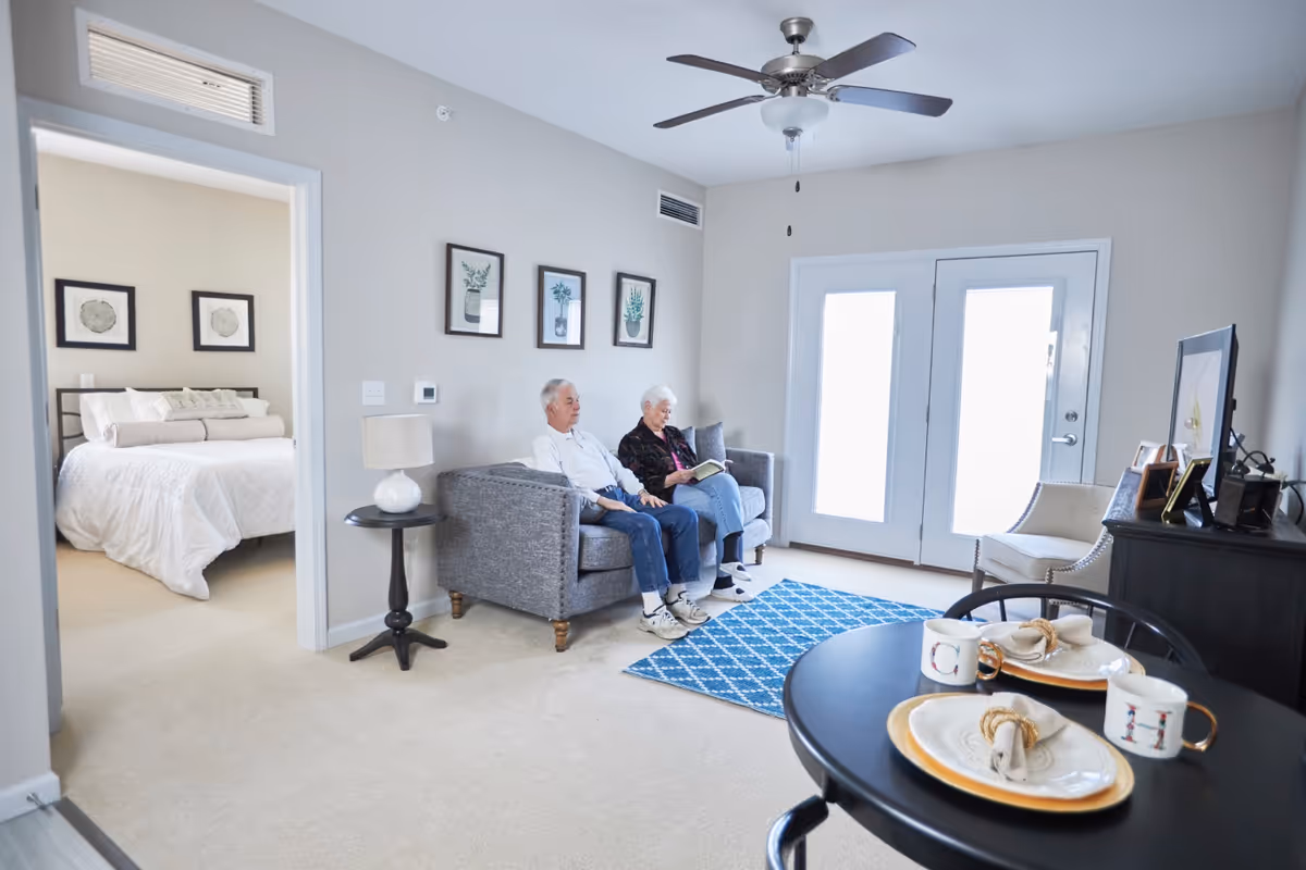 A cozy senior living room with an elderly couple sitting on a gray couch. The room features a ceiling fan, a blue patterned rug, and a small round dining table set with plates and mugs. A doorway leads to a bedroom with a neatly made bed and framed artwork on the walls. French doors allow natural light to brighten the space.