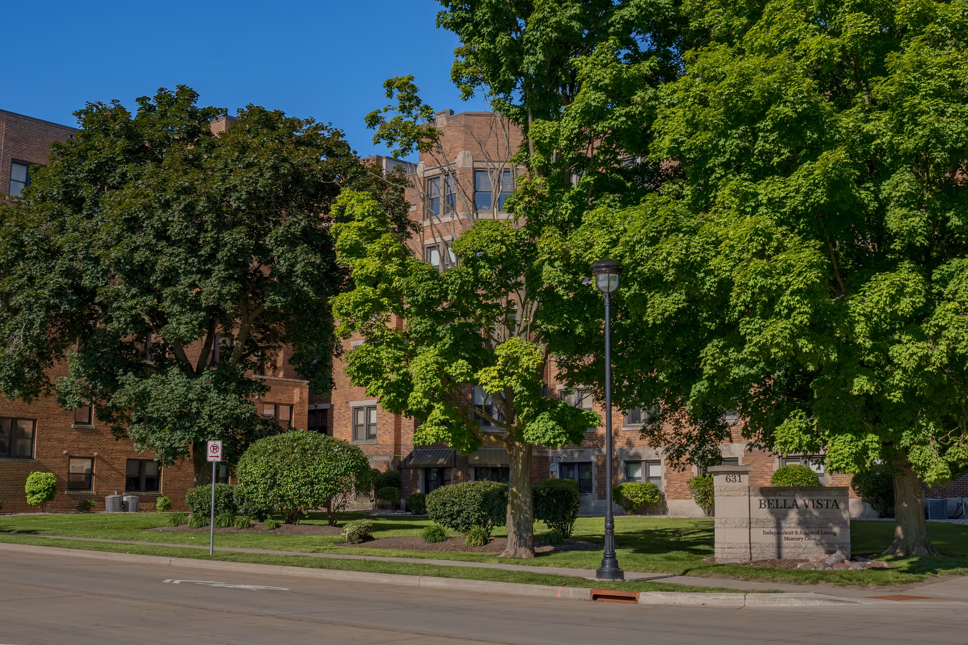 Exterior view of the Bella Vista senior living facility showing a multi-story brick building partially obscured by large green trees and landscaping. A stone sign near the sidewalk reads 'Bella Vista' with additional text about independent and assisted living and memory care. A street lamp and a no parking sign are also visible.