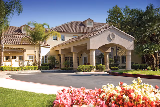 Exterior view of a senior living facility named Park Terrace with a beige stucco building, tiled roof, arched entrance, palm trees, and colorful flowers in the foreground under a clear blue sky.