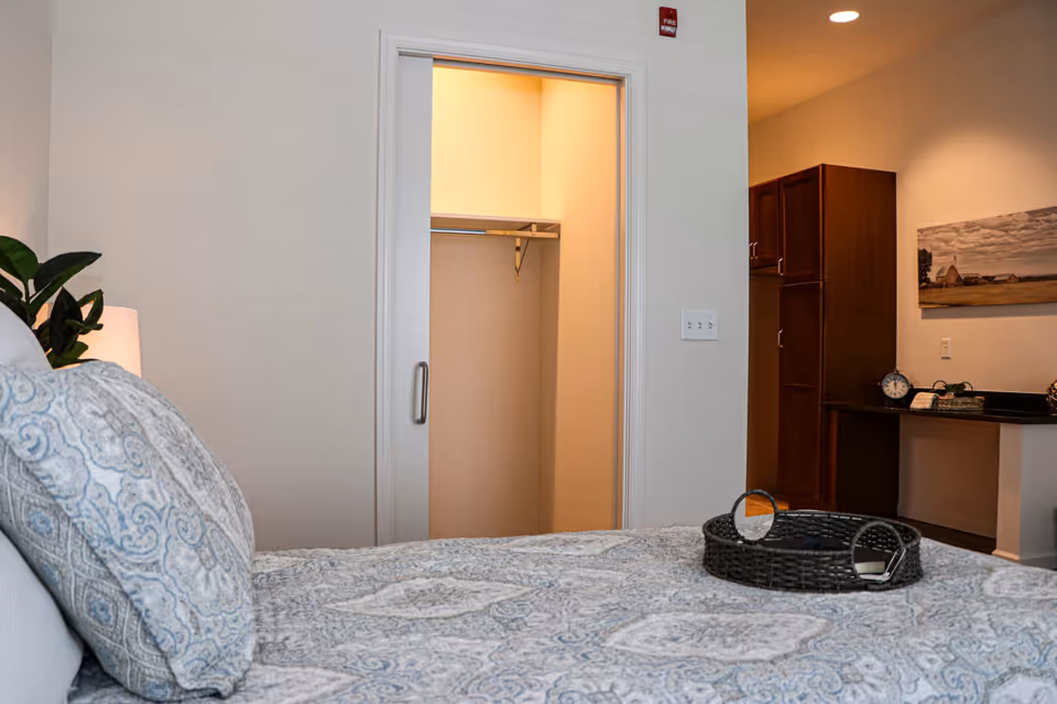 A bedroom with a neatly made bed featuring patterned pillows and a matching bedspread. There is a small plant and lamp on the left side. In the background, an open closet with a hanging rod is visible, along with a wooden cabinet and a desk area with a clock and decorative items. A landscape painting hangs on the wall above the desk.