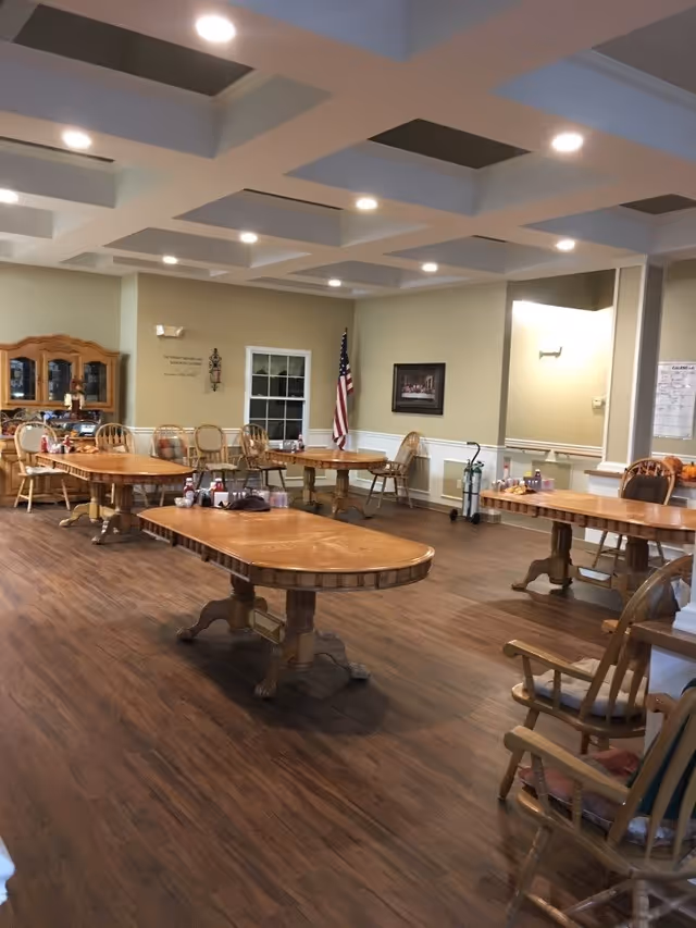 A spacious dining room with several wooden tables and chairs arranged neatly. The room has a wooden floor, beige walls with white wainscoting, a ceiling with recessed lighting, and an American flag in the corner. There is a wooden cabinet with glass doors against one wall and a framed picture hanging on another wall.