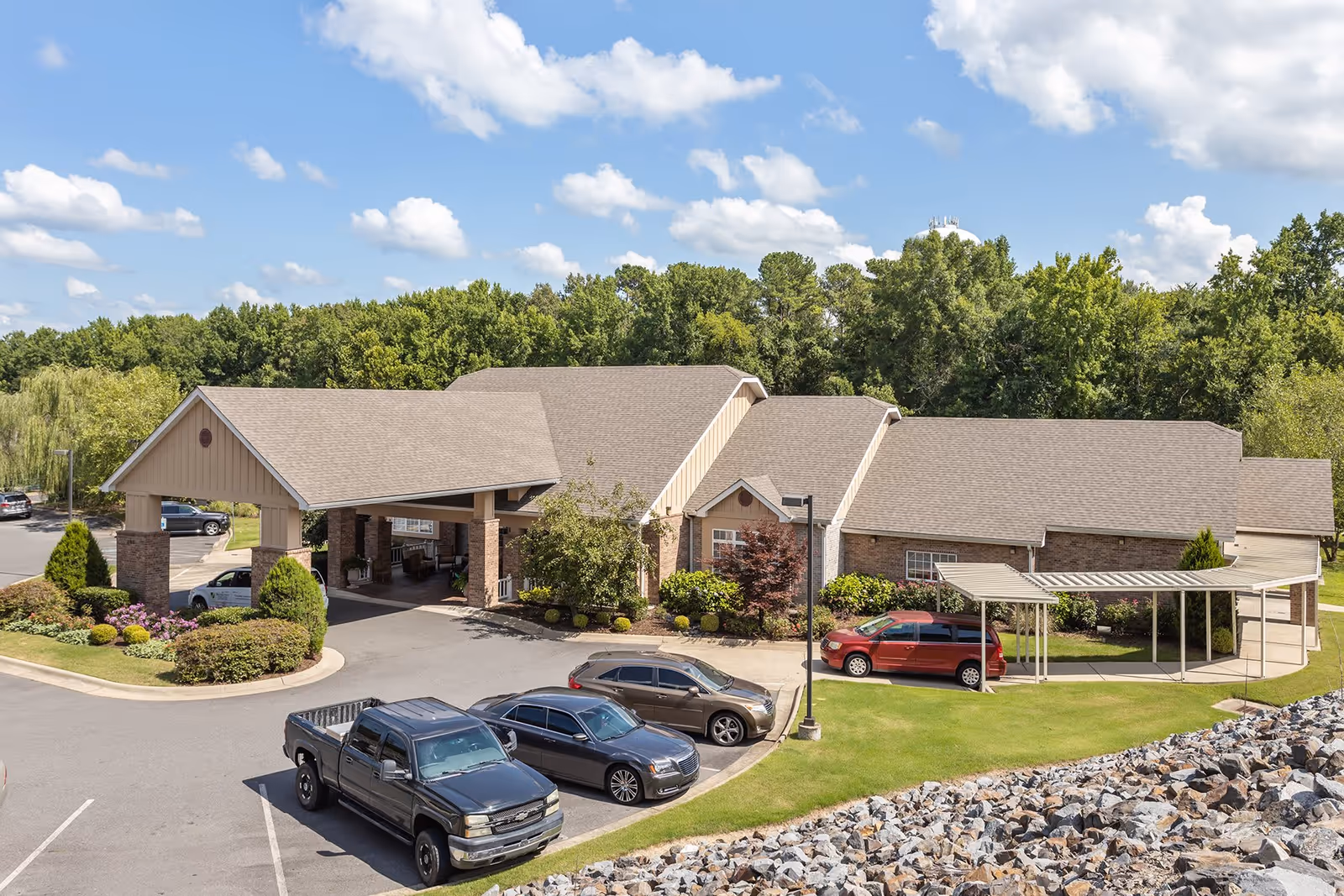 Exterior view of Brookfield Assisted Living and Memory Care facility showing a single-story building with a covered entrance, several parked cars, landscaped greenery, and a backdrop of trees under a partly cloudy sky.