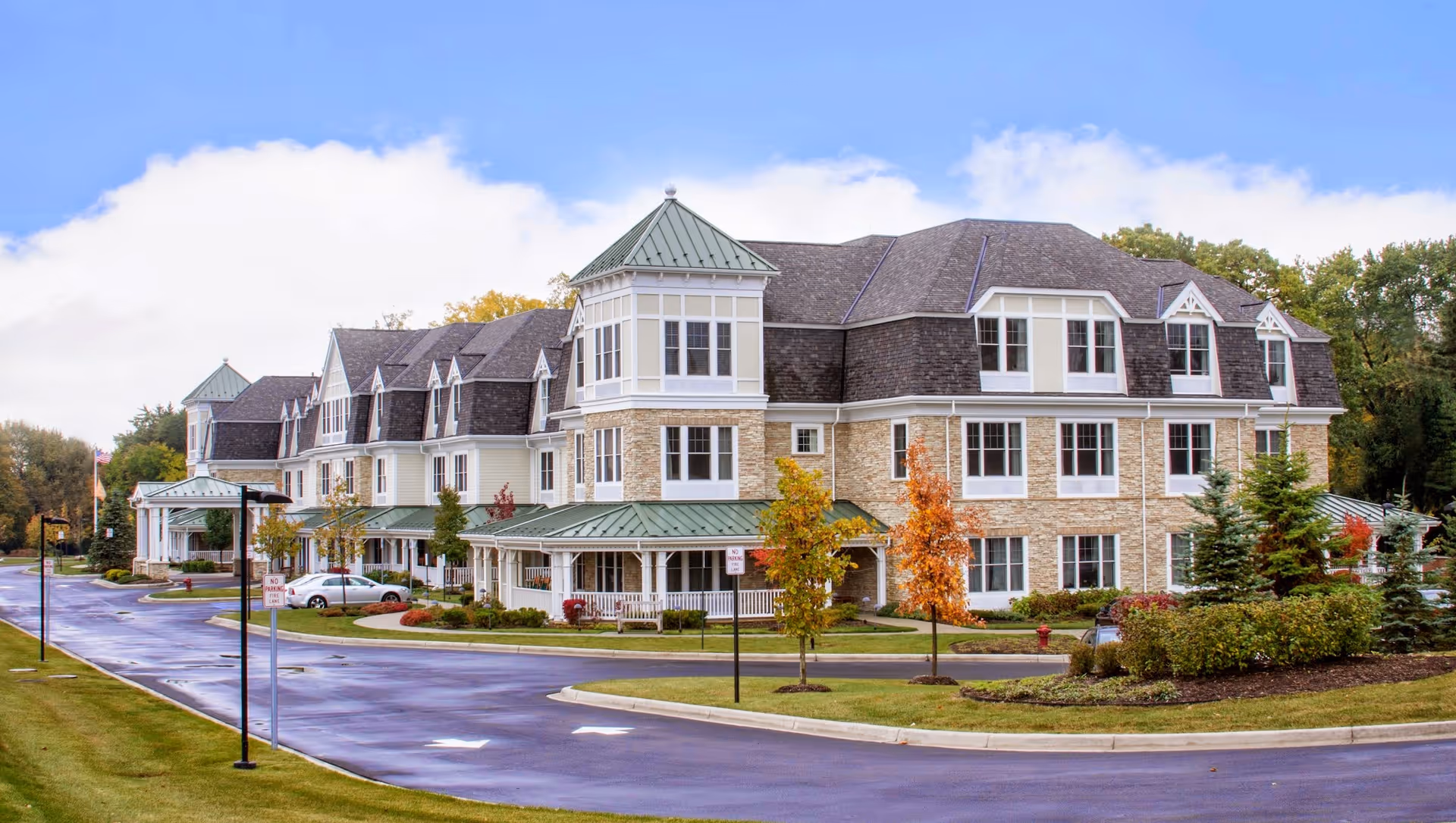 Exterior view of a large, multi-story senior living facility building with a combination of stone and siding facade, green metal roofs, and multiple windows. The building is surrounded by landscaped greenery, small trees with autumn foliage, and a paved driveway with street lamps.