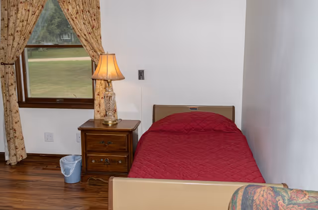 Small bedroom with a single bed covered in a red quilt, wooden nightstand with a lamp, window with floral curtains, and hardwood floor.