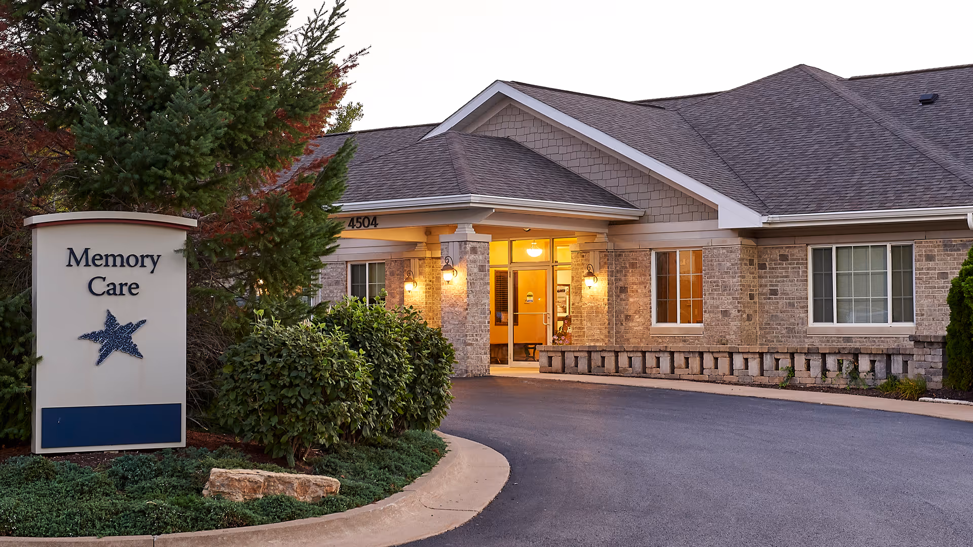 Exterior view of a senior living facility entrance with a sign that reads 'Memory Care' next to a driveway. The building has brick walls, lit wall lamps, and large windows.