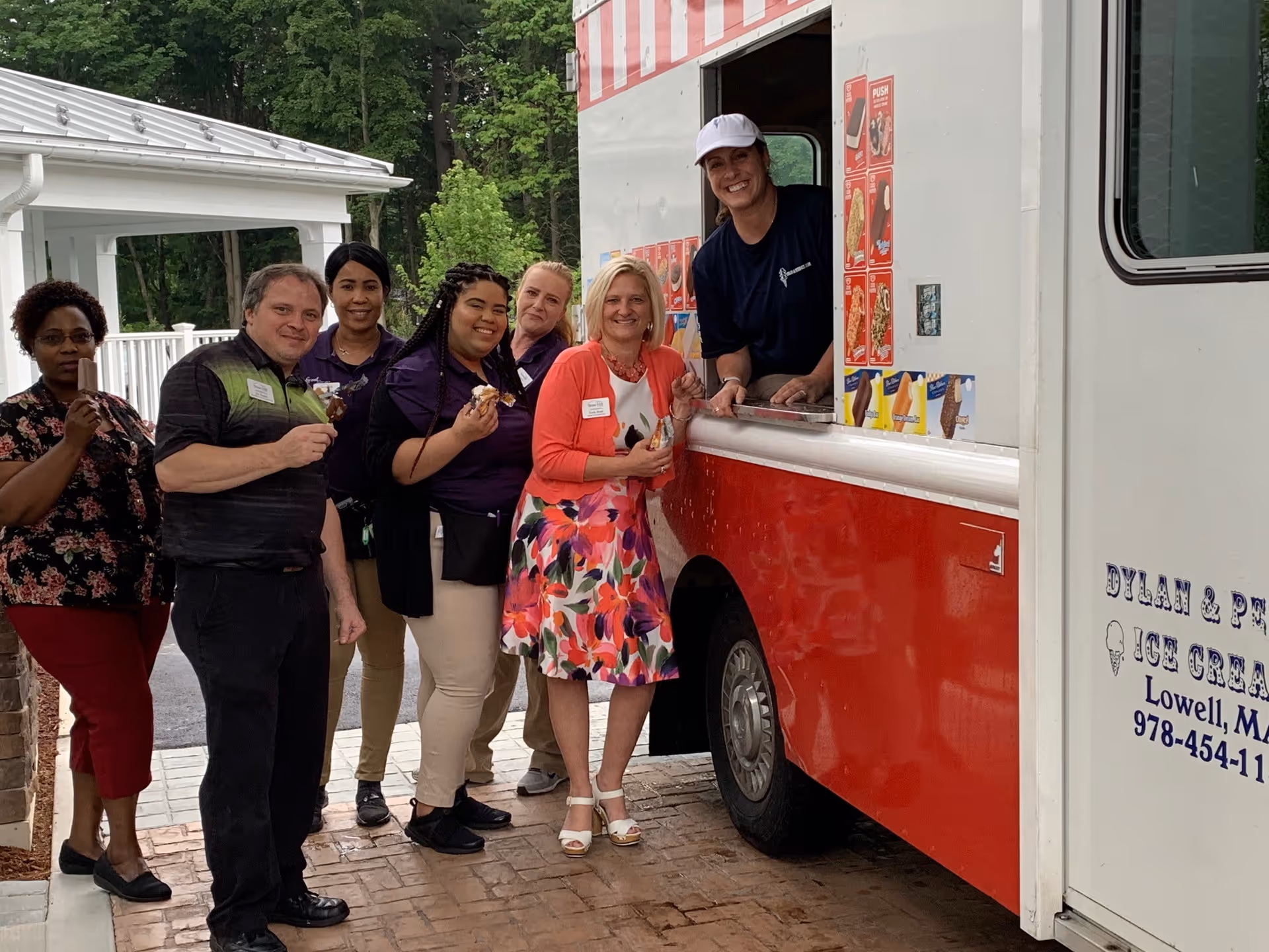 A group of six adults standing outside next to a red and white ice cream truck. Five people are standing in front of the truck holding ice cream treats, smiling at the camera. One person is inside the truck serving ice cream. The background shows greenery and part of a white building.