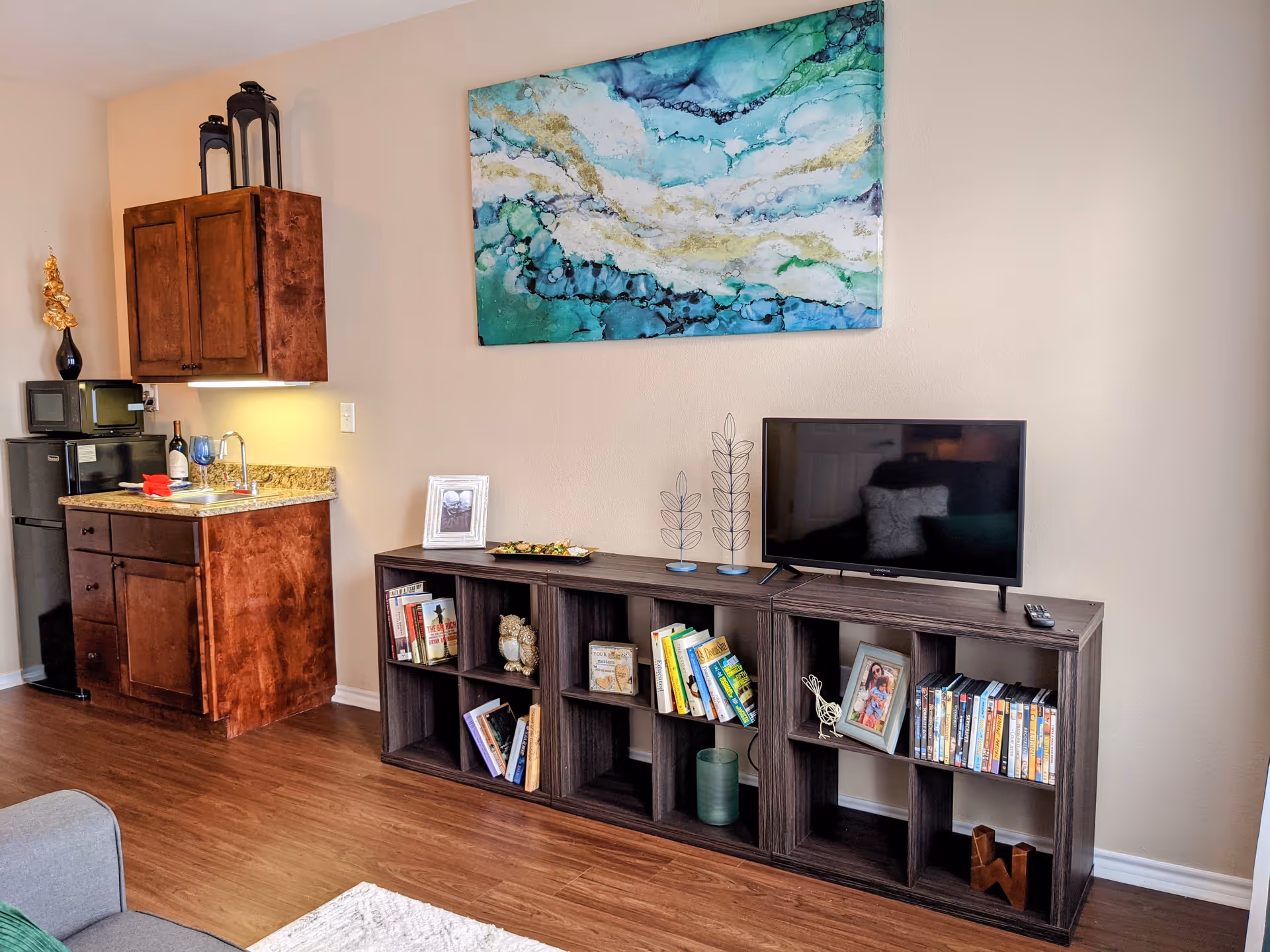 Interior view of a living space featuring a small kitchenette with a mini fridge, microwave, and wooden cabinets on the left. To the right, there is a dark wooden shelving unit with books, DVDs, framed photos, and decorative items. A flat-screen TV is placed on top of the shelving unit. Above the TV, a large abstract painting with blue, green, and white colors hangs on the wall. The floor is wooden, and part of a gray couch and a white rug are visible in the foreground.