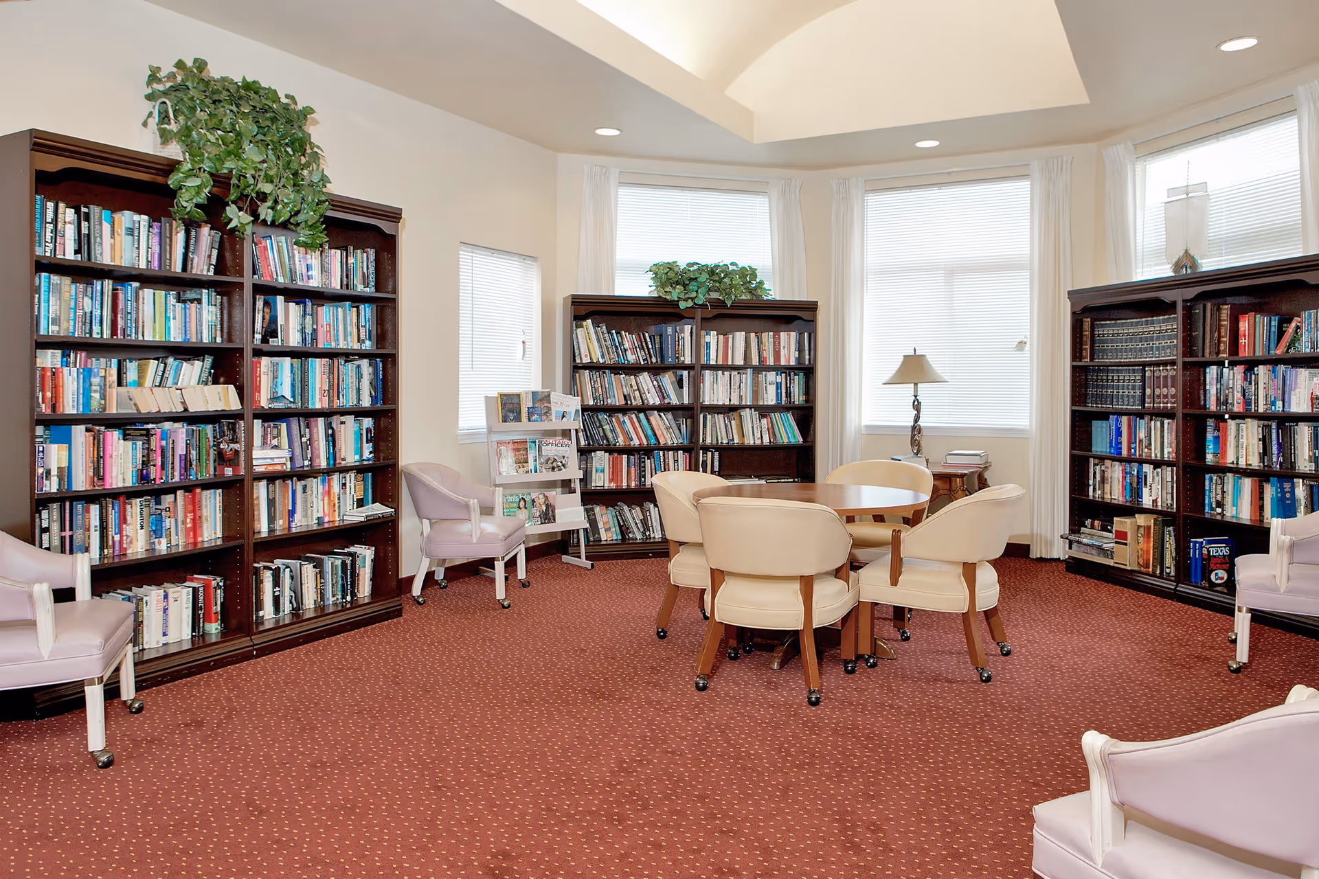 A cozy library room with three large wooden bookshelves filled with books, a round wooden table surrounded by four beige chairs, and several lavender armchairs on wheels. The room has a red carpet with a subtle pattern, large windows with white blinds and curtains, and a ceiling with recessed lighting and a skylight. There are green plants on top of the bookshelves and a small lamp on a side table near the windows.