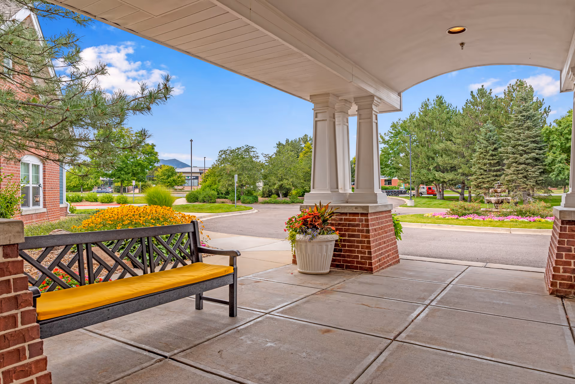 Covered outdoor entrance area with a black bench featuring a yellow cushion, brick pillars, a large planter with colorful flowers, and a view of a landscaped driveway with trees, bushes, and a fountain in the background under a blue sky with some clouds.