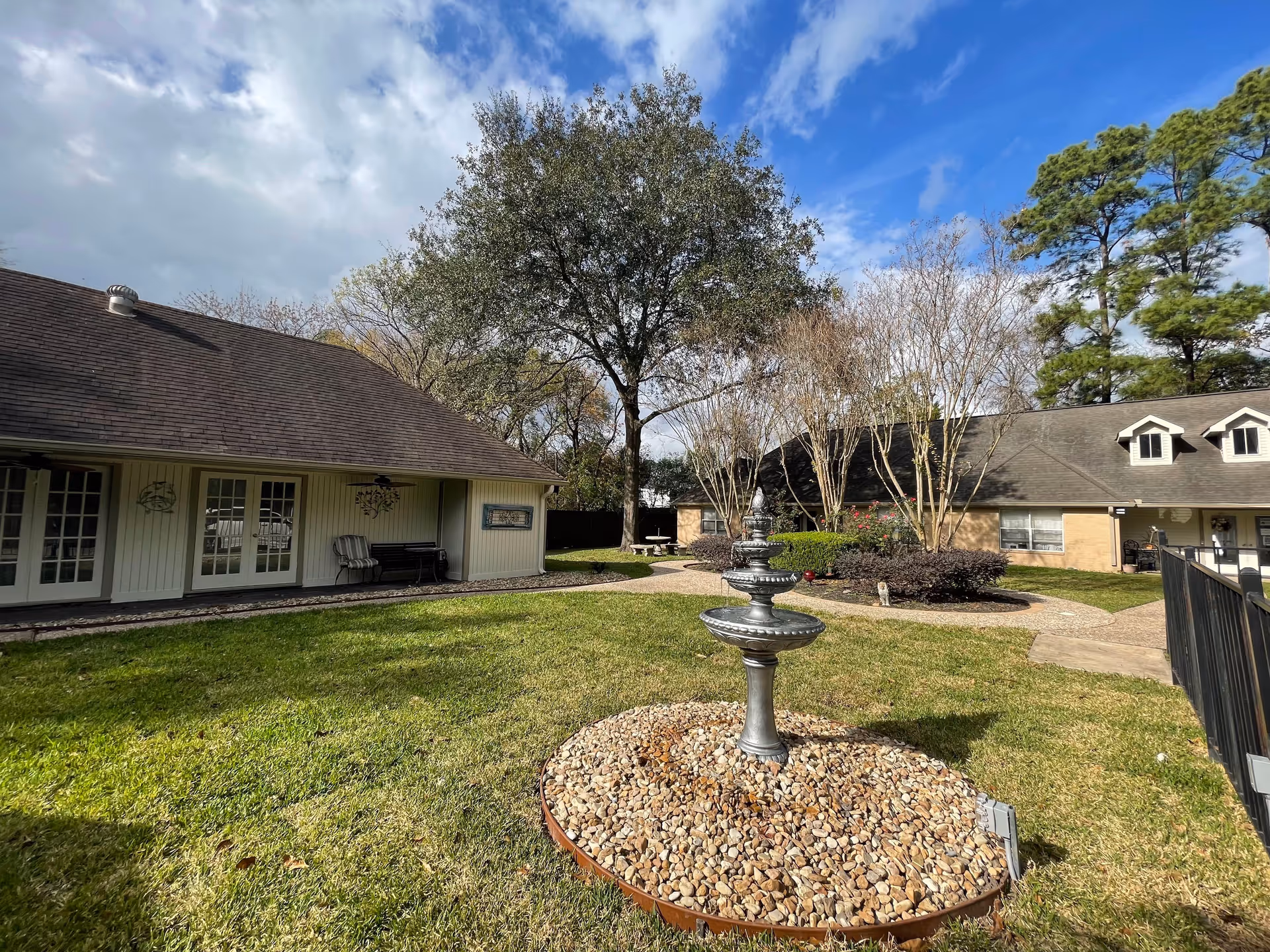 Outdoor garden area of Tranquility Personal Care Home featuring a central decorative fountain surrounded by small rocks, green grass, trees, and two buildings with multiple windows and doors under a partly cloudy sky.