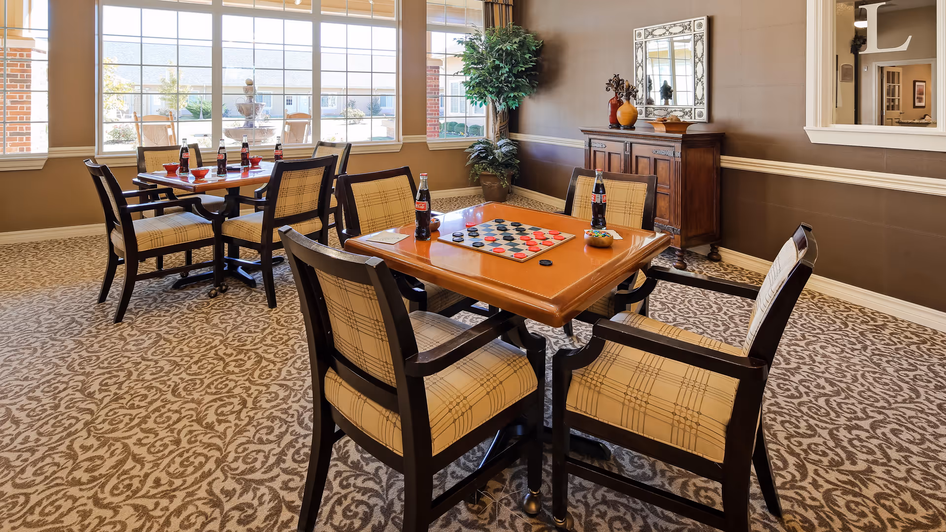 A well-lit common area with two wooden tables surrounded by cushioned chairs. One table has a checkers board set up with red and black pieces, and several bottles of Coca-Cola are placed on both tables. The room features large windows letting in natural light, a patterned carpet, a wooden sideboard with decorative items, a large mirror on the wall, and a potted plant in the corner.