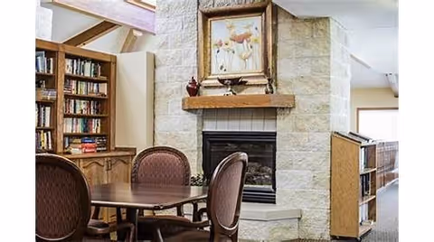 Cozy common room featuring a stone fireplace topped with framed artwork, a round table with chairs, and bookshelves.