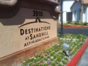 A close-up view of a beige stone sign with the text 'Destinations at Sandhill Active Adult Living' and the number 3210 above it, surrounded by a landscaped area with small plants and flowers, with a building and driveway visible in the background.