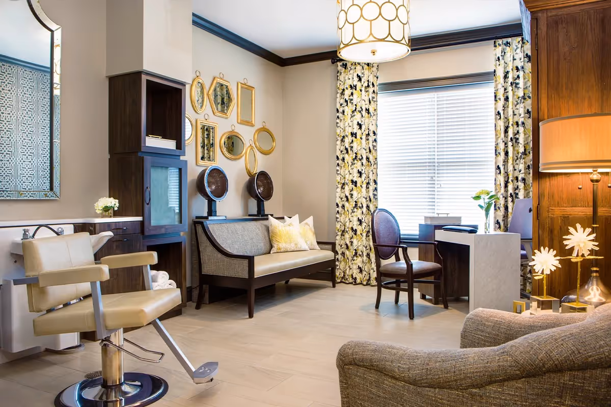 A stylish interior room with a beige salon chair in front of a small sink, a wooden bench with cushions, decorative gold-framed mirrors on the wall, a desk with a chair near a window with floral curtains, and a floor lamp with a beige shade. The room has light-colored flooring and a cozy, welcoming atmosphere.