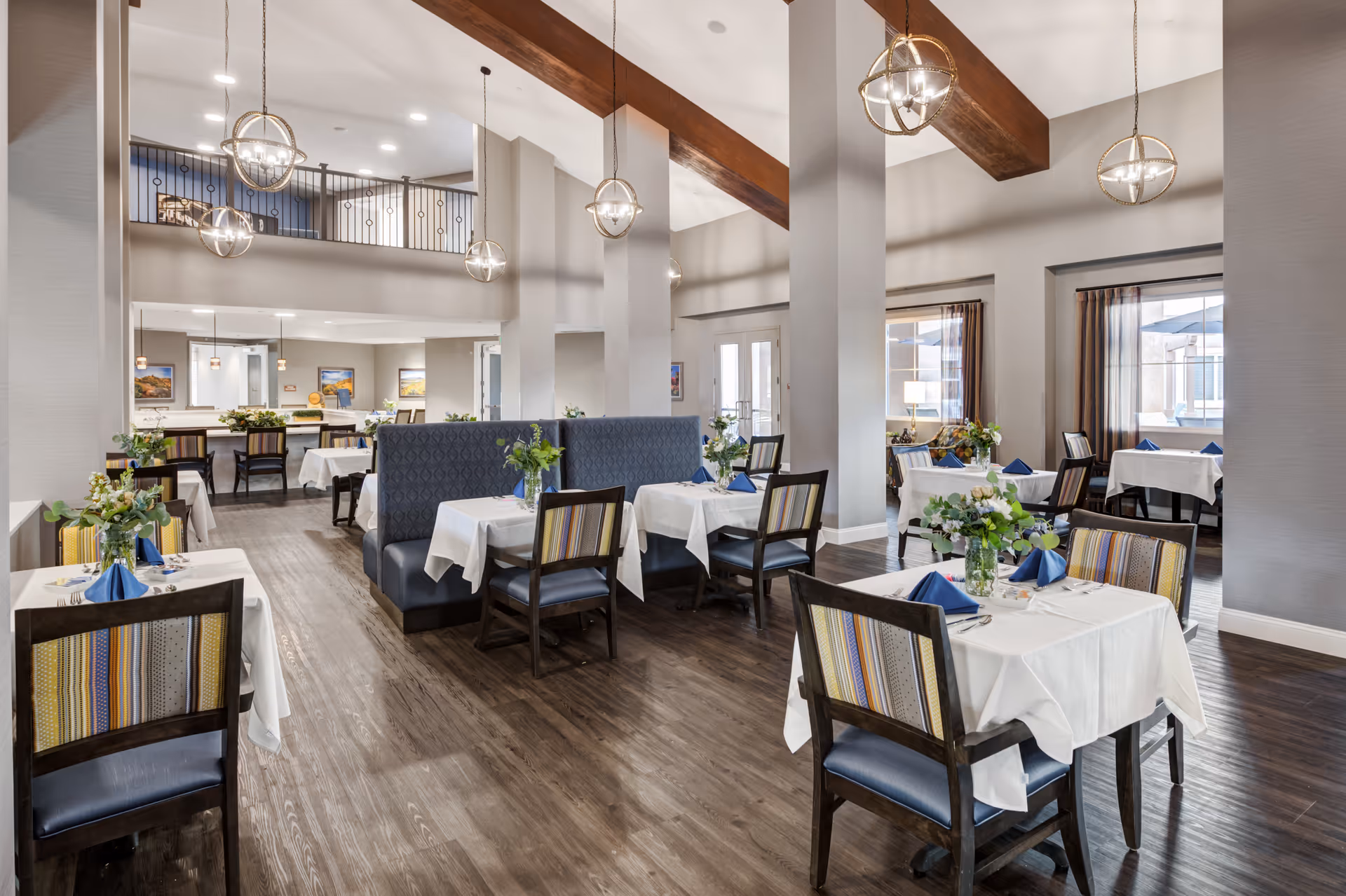 Bright upscale dining room with set tables, floral centerpieces, blue napkins, high ceilings with wooden beams and globe pendant lights.