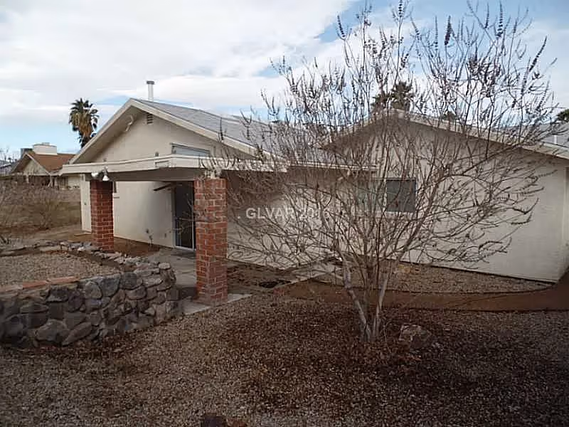 Exterior view of a single-story building with a covered entrance supported by brick pillars. The surrounding yard has gravel ground cover and a leafless tree in the foreground. The sky is partly cloudy.