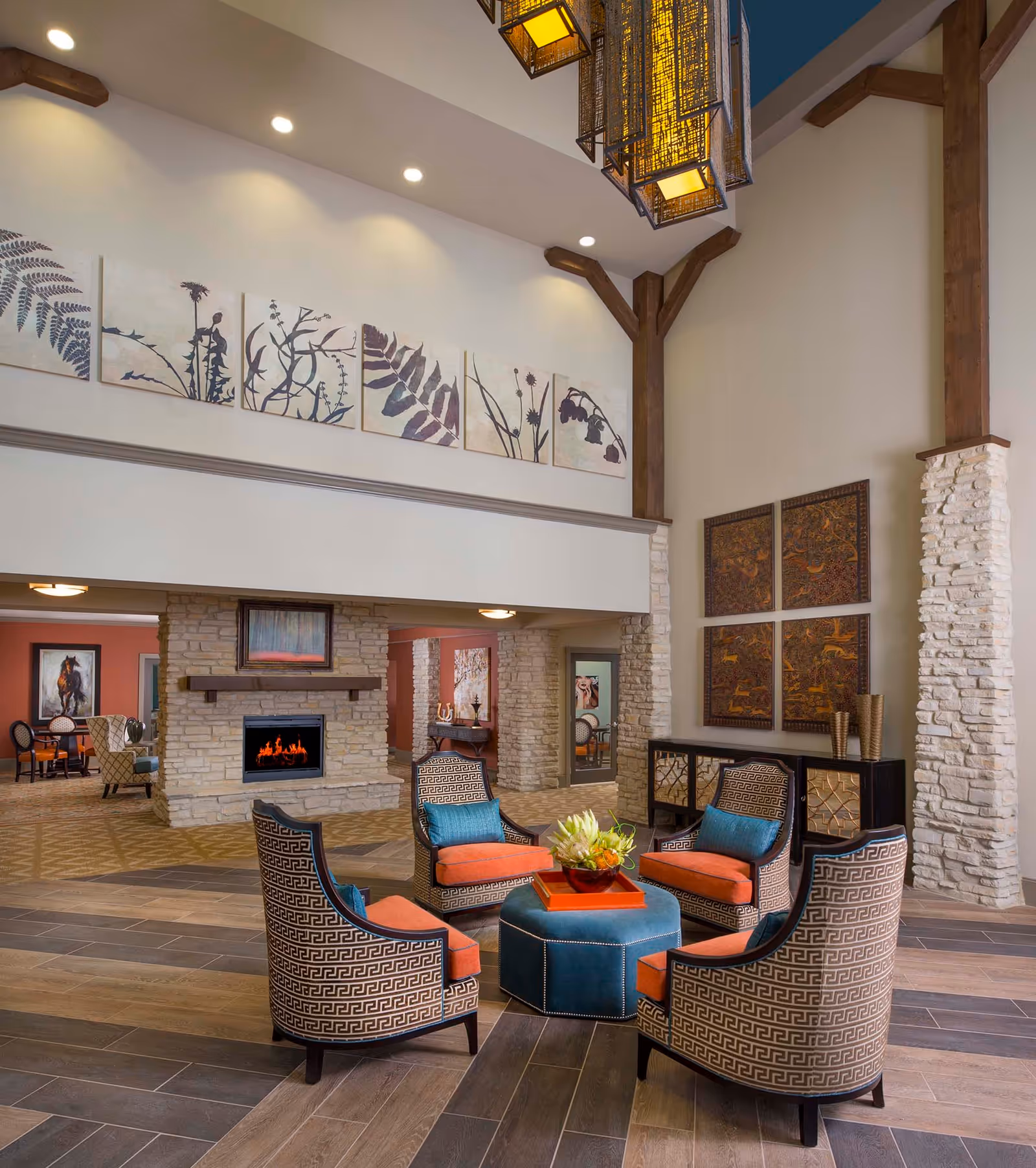 A spacious and well-lit living room area in an assisted living facility featuring a circular arrangement of five patterned armchairs with orange cushions and teal pillows around a blue ottoman with a decorative centerpiece. The room has a high ceiling with wooden beams, a stone fireplace with a fire burning, and various pieces of artwork on the walls. The flooring is a mix of wood and tile, and there is a dark wooden console table with decorative vases against one wall.