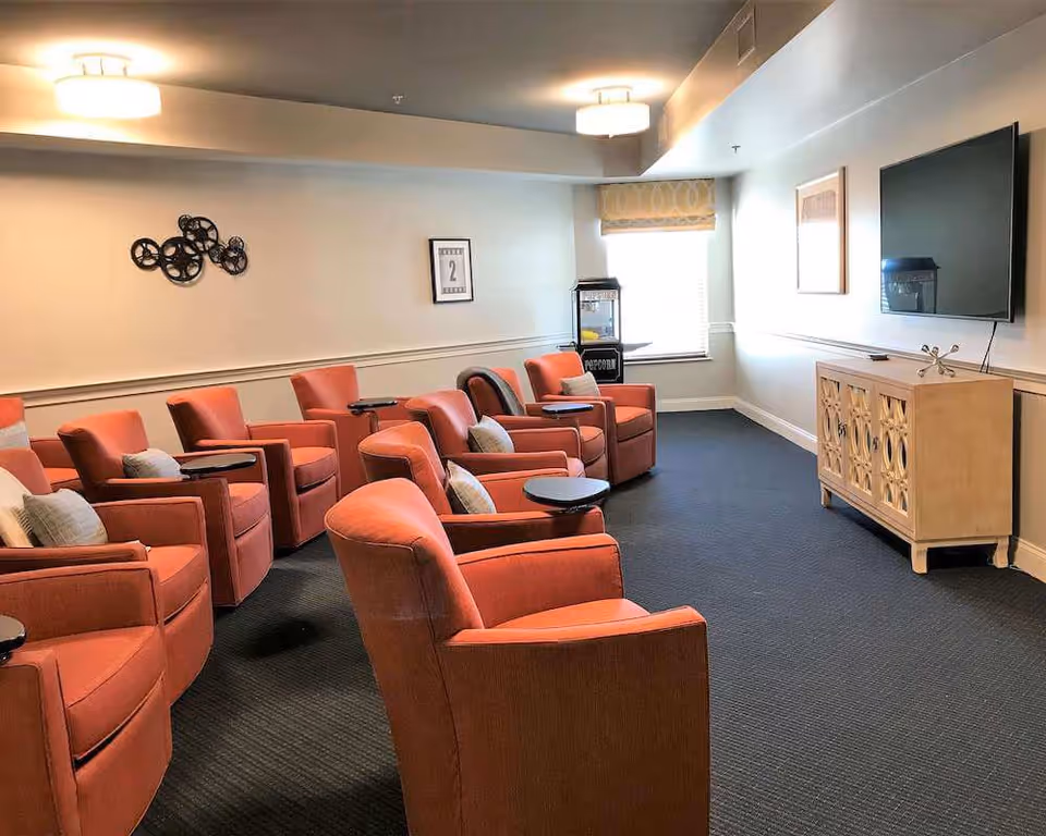 Small media room with rows of orange armchairs facing a wall-mounted TV and a popcorn machine by a window.